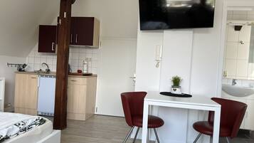 A modern room with a bed, a dining table with two chairs, a TV mounted on the wall, a kitchen area with cabinets and a fridge, and a bathroom with a sink and mirror.