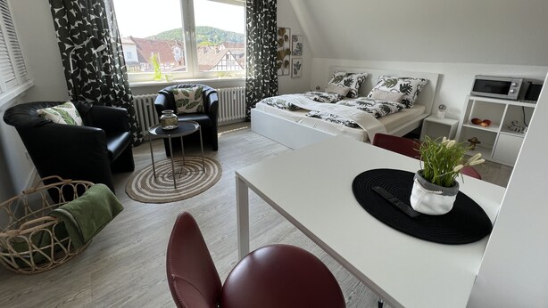 A modern, well-lit room with a bed, a desk, a chair, and a view of a green landscape through large windows. - Marketplace apartment (Höxter)