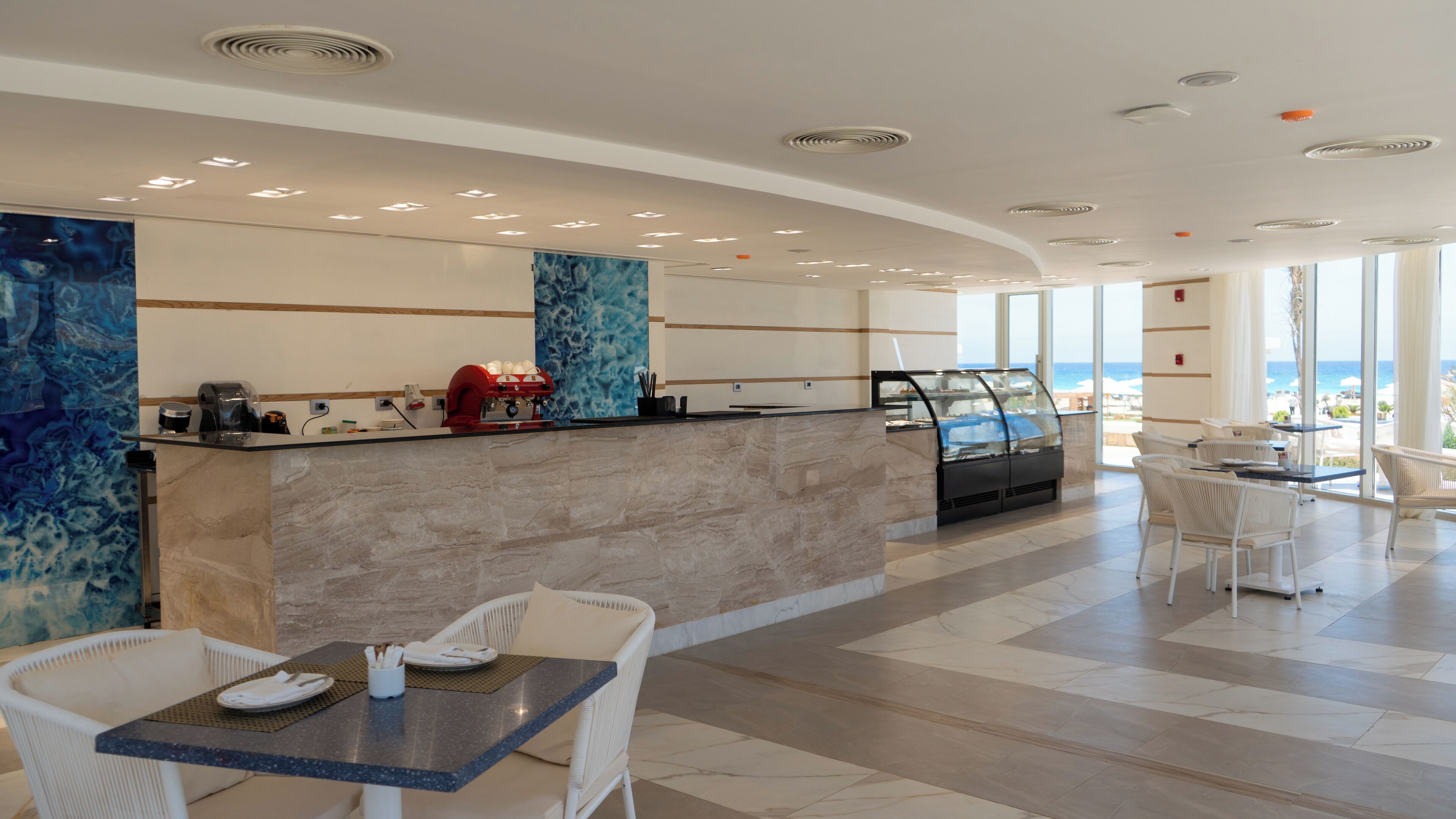 A modern hotel lobby with a marble reception desk, seating area, and large windows offering a view of the beach.