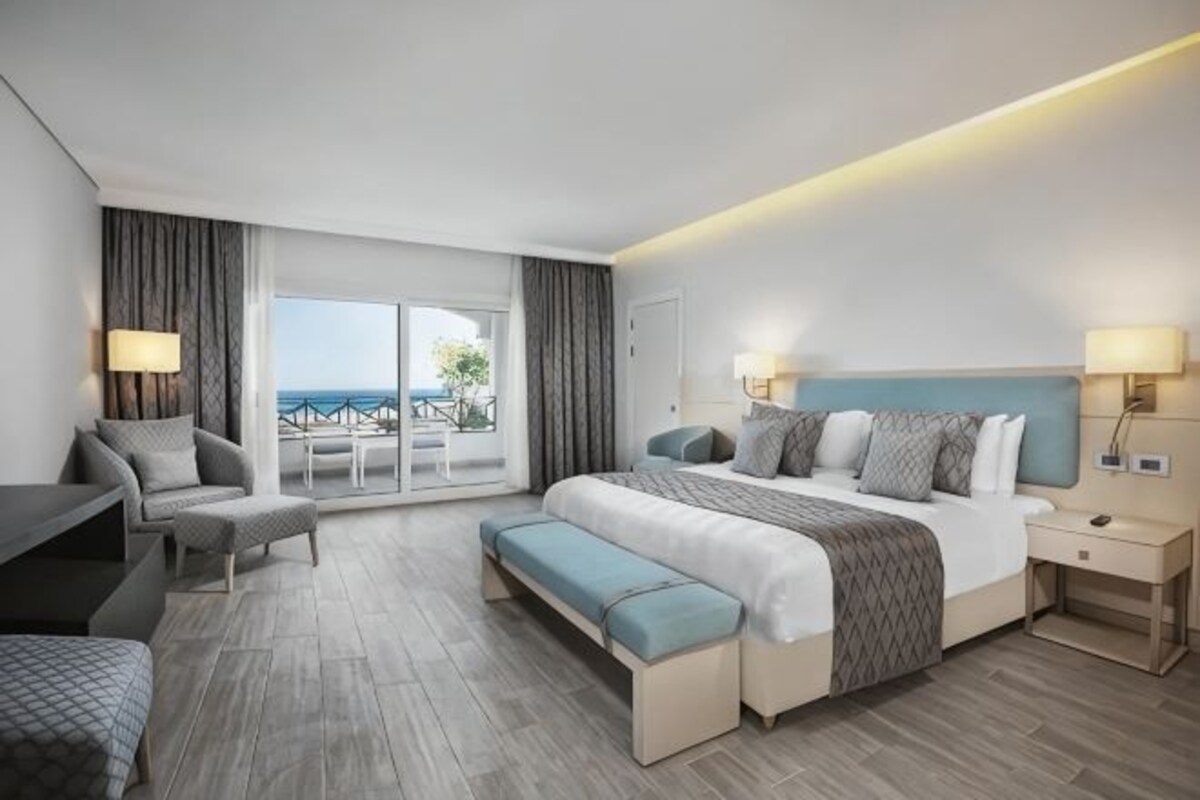 A modern hotel room with a large bed, a seating area, a desk, and a balcony with a view.