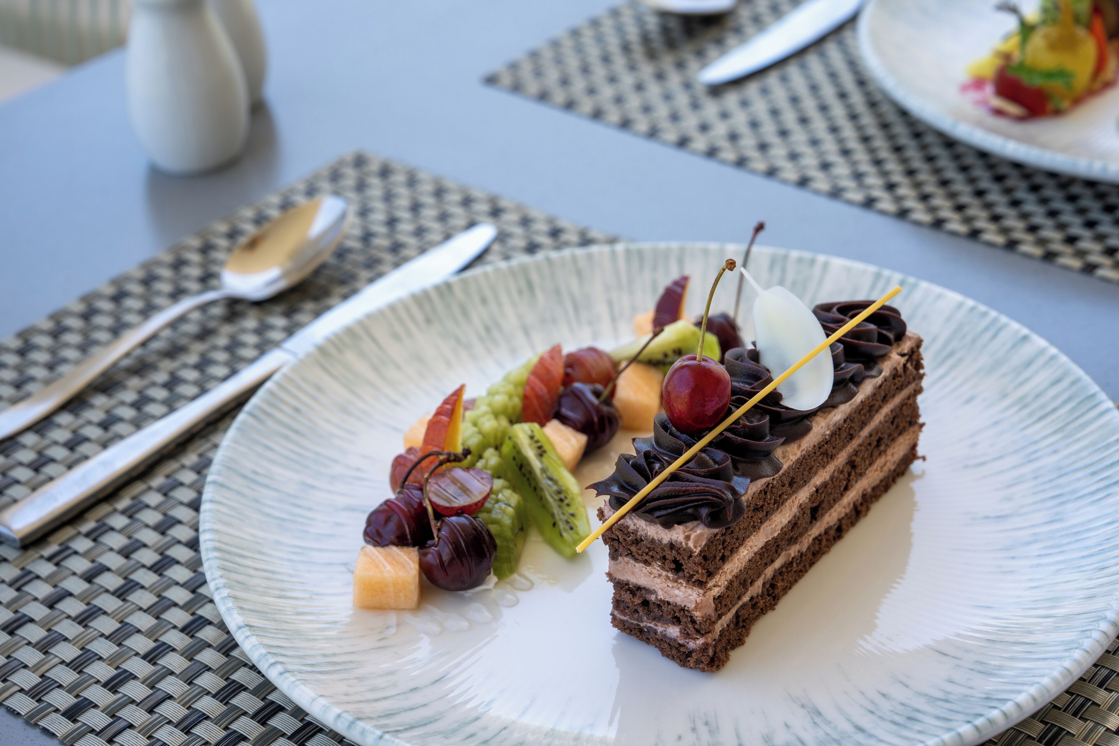 A dessert plate with chocolate cake, cherries, kiwi, and grapes on a woven placemat.