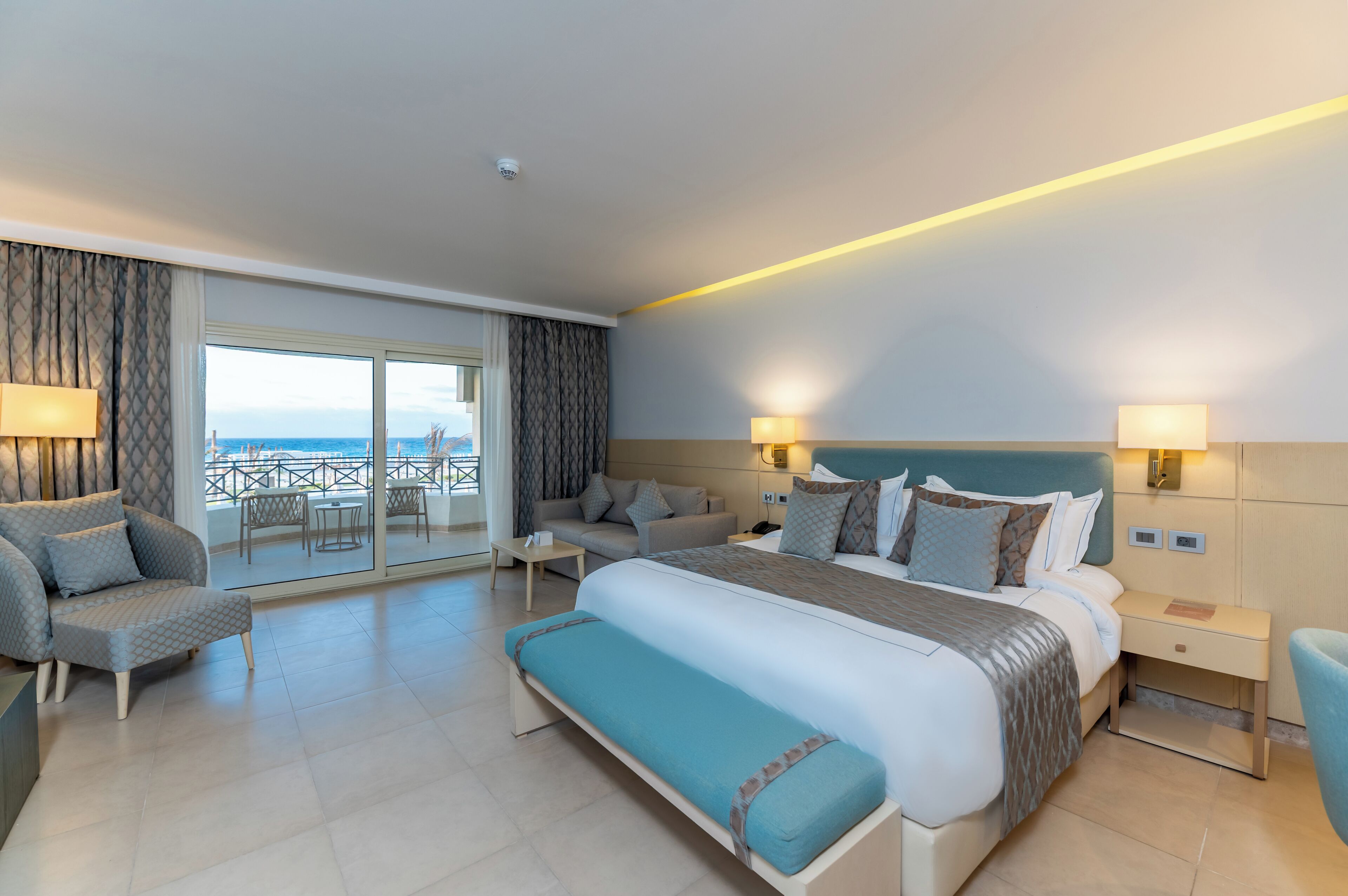 A modern hotel room with a large bed, two bedside tables, a sofa, a bench, and a balcony with a view of the sea.