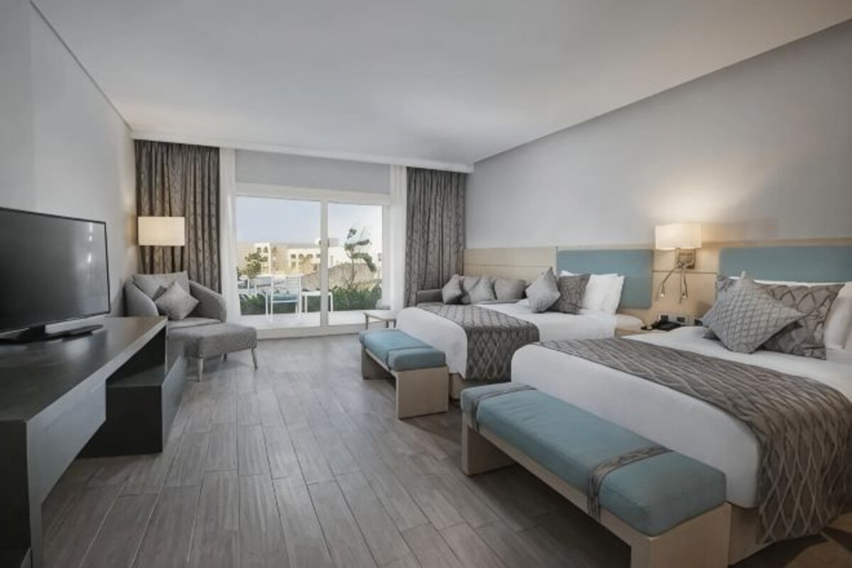 A hotel room with a large bed, a smaller bed, a desk with a TV, and a balcony with curtains.
