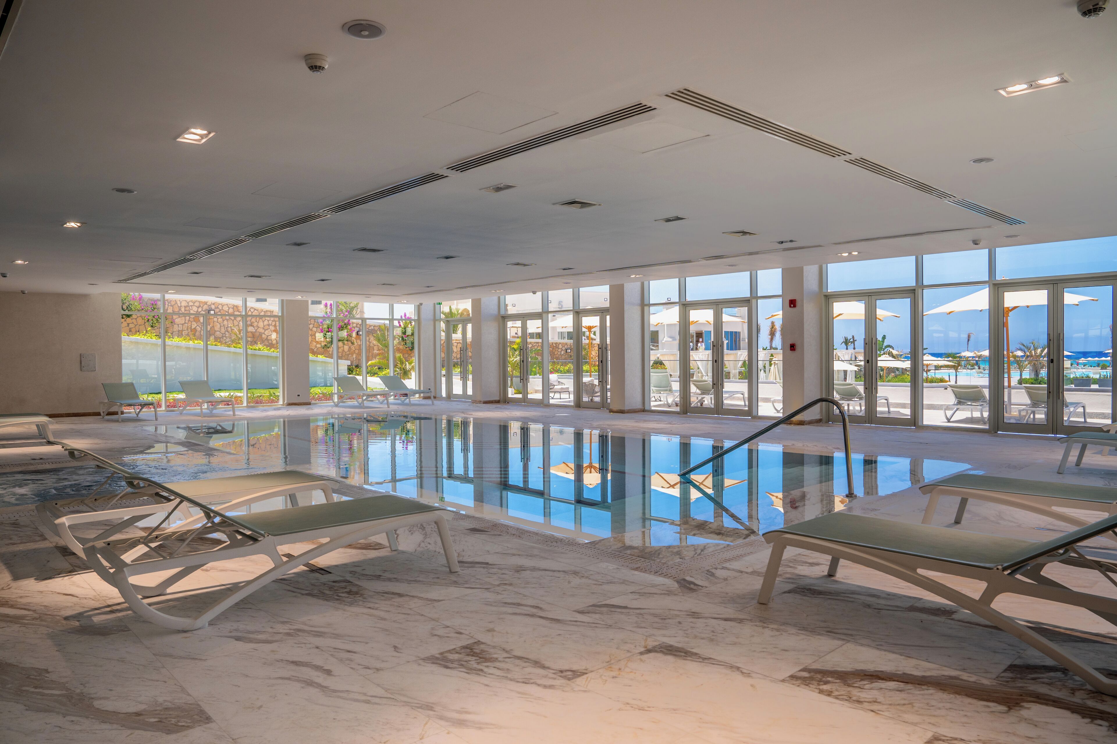 Une piscine intérieure avec des parois en verre transparent, des chaises longues et une vue sur l’extérieur.