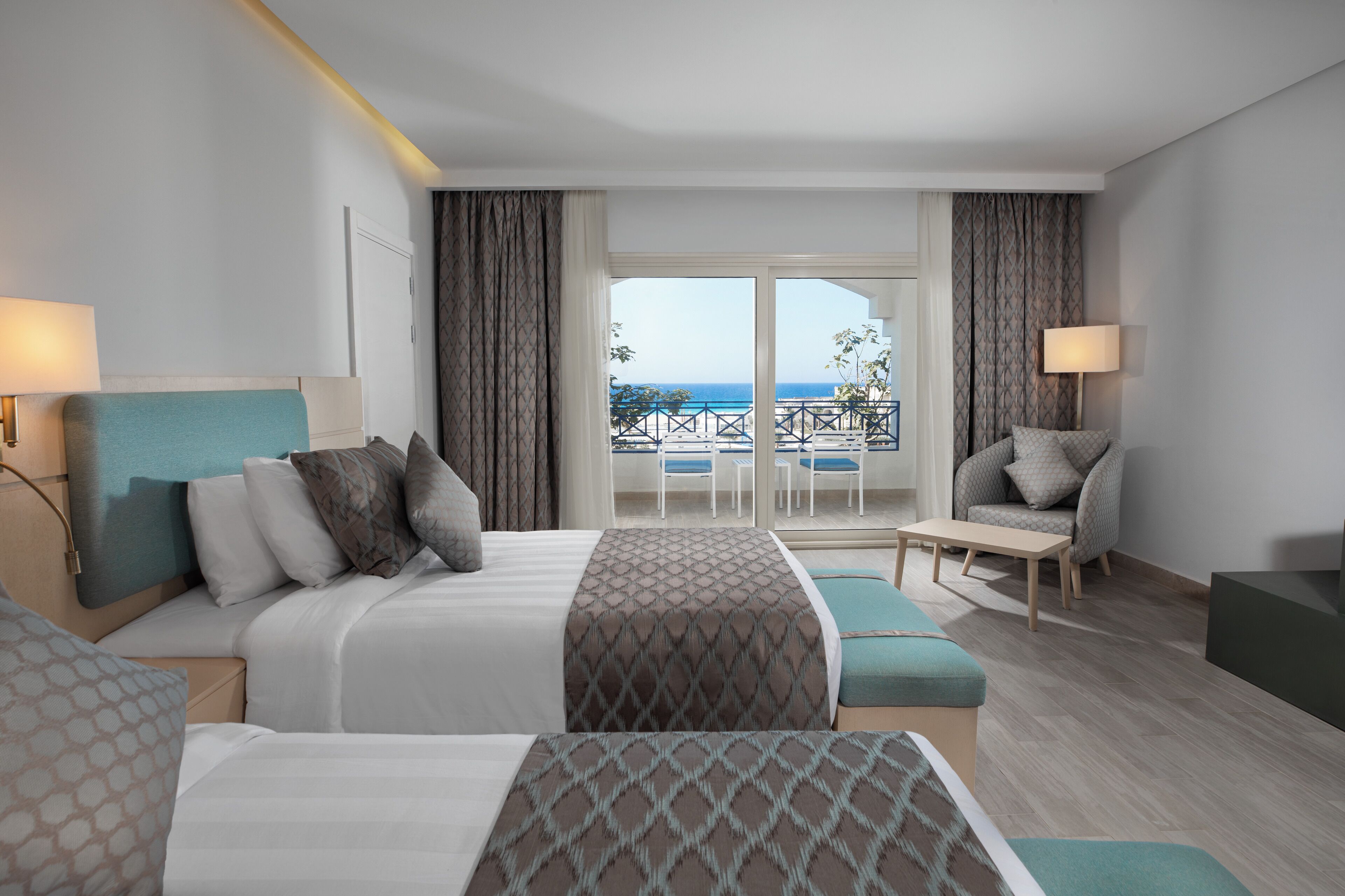 A hotel room with a large bed, a sofa, a small table, and a balcony with a view of the sea.