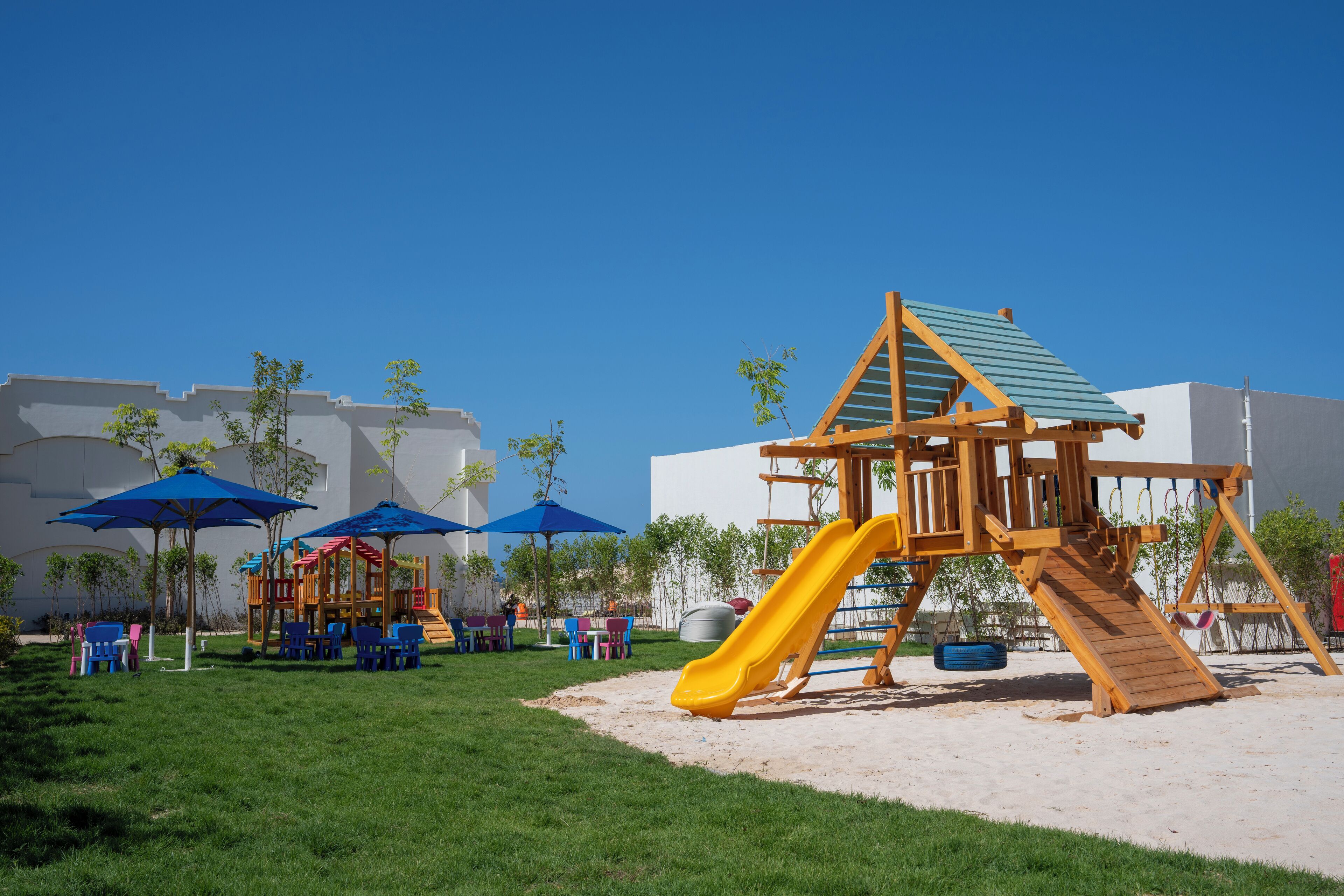 Un parque infantil con tobogán y estructura de escalada, rodeado de césped verde y una zona de arena.
