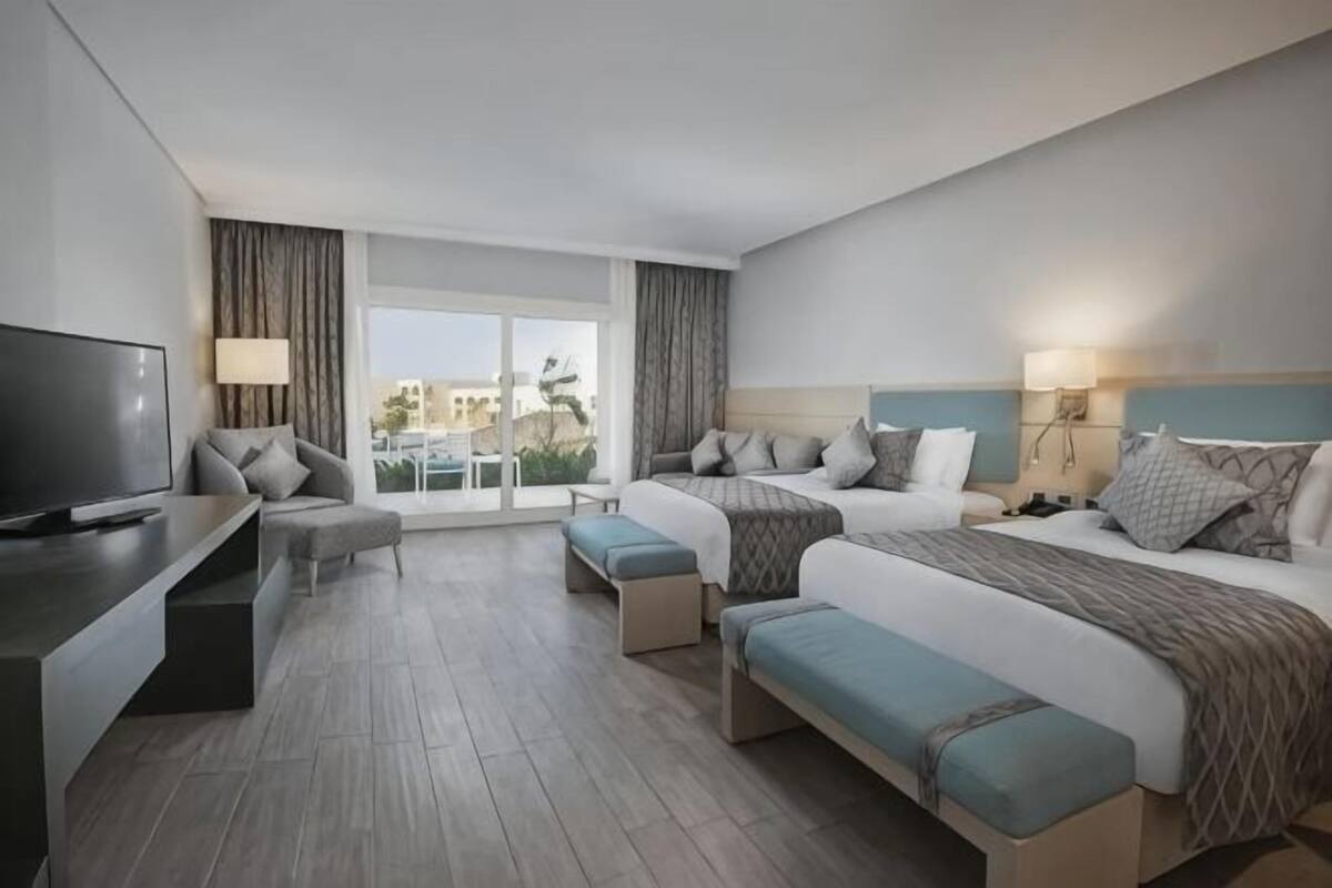Habitación de hotel con una cama grande, una cama más pequeña, un escritorio con televisor y un balcón con cortinas.