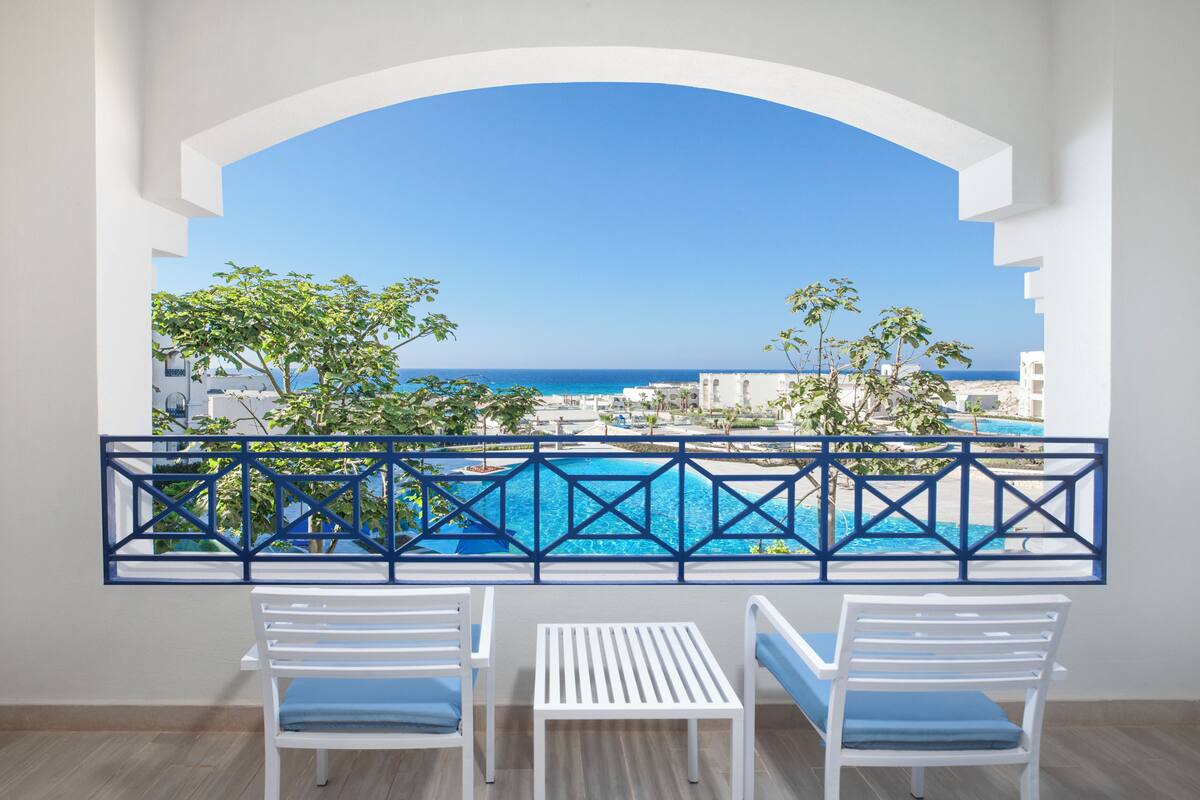 Balcón con vistas a una piscina y al mar, con sillas blancas y barandilla azul.