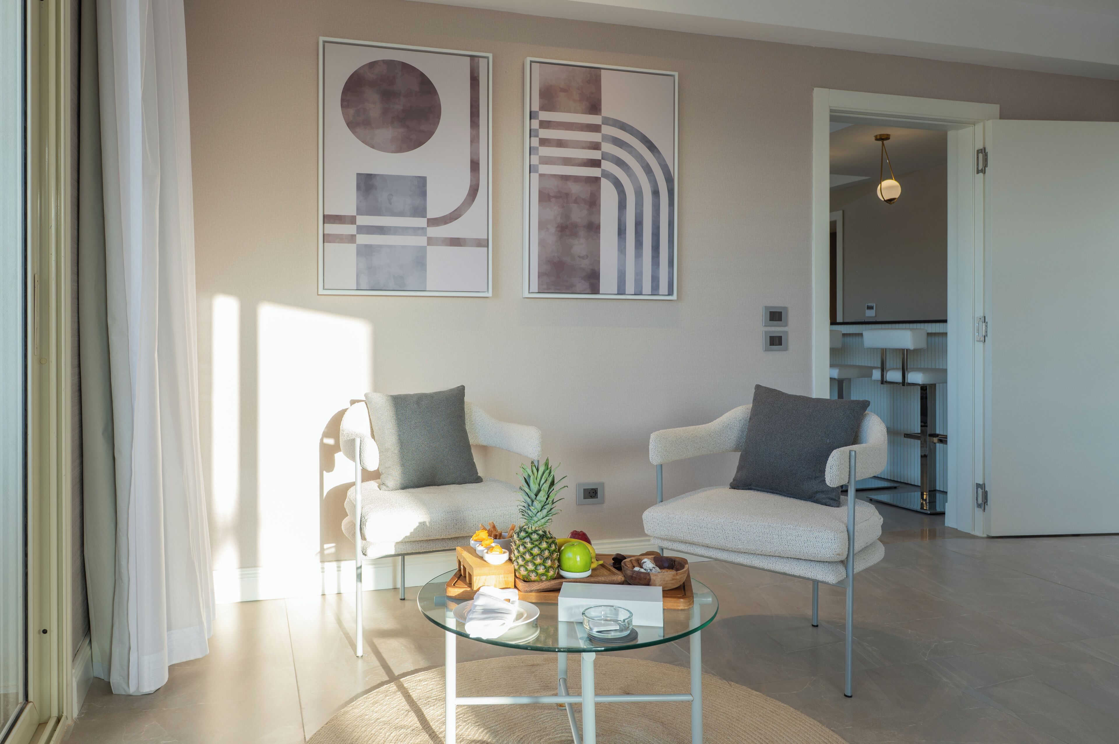 Una sala de estar moderna con dos sillones blancos, una mesa de centro de cristal con frutas y aperitivos, y una obra de arte abstracto en la pared.
