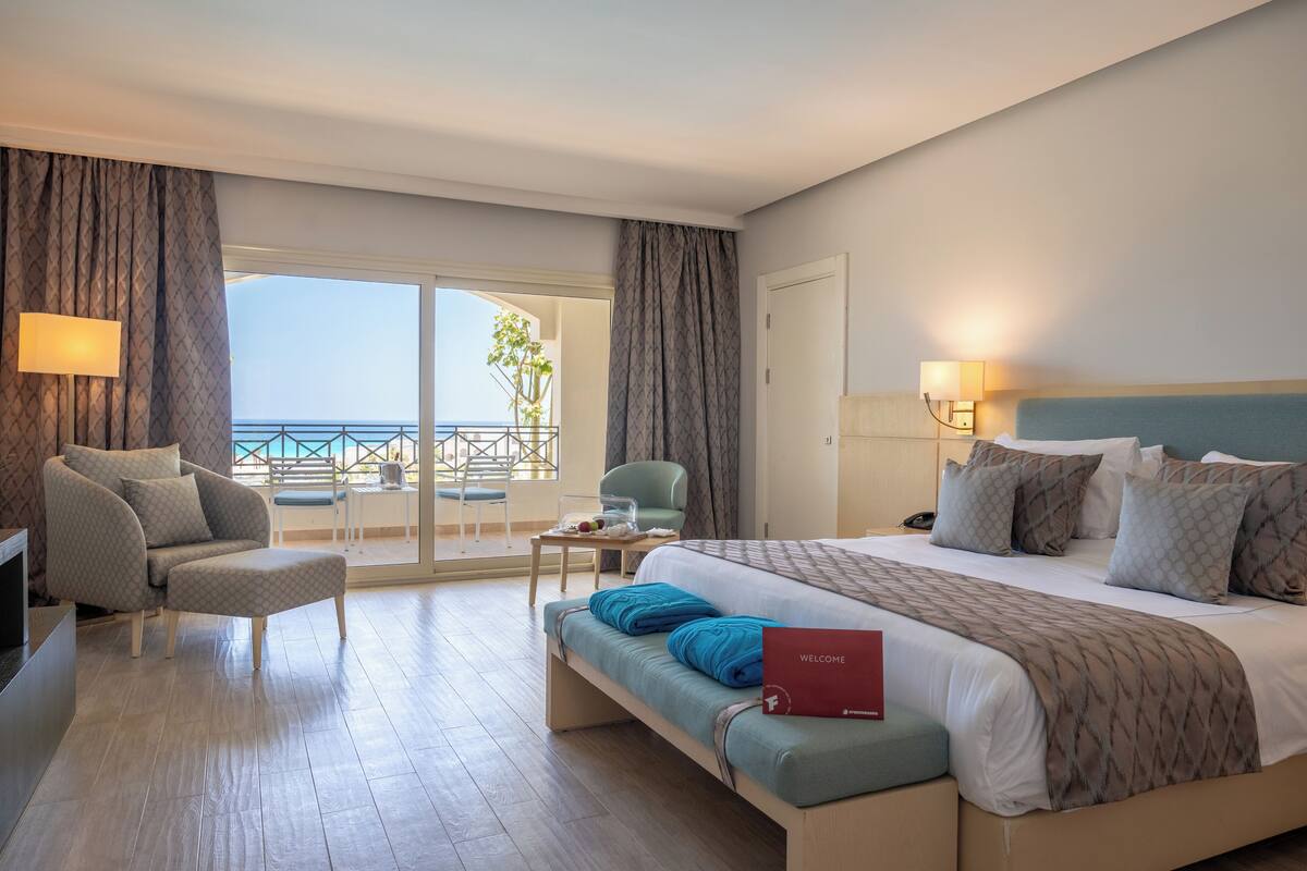 Habitación de hotel con cama, sofá, sillón, una mesita y vistas al mar.