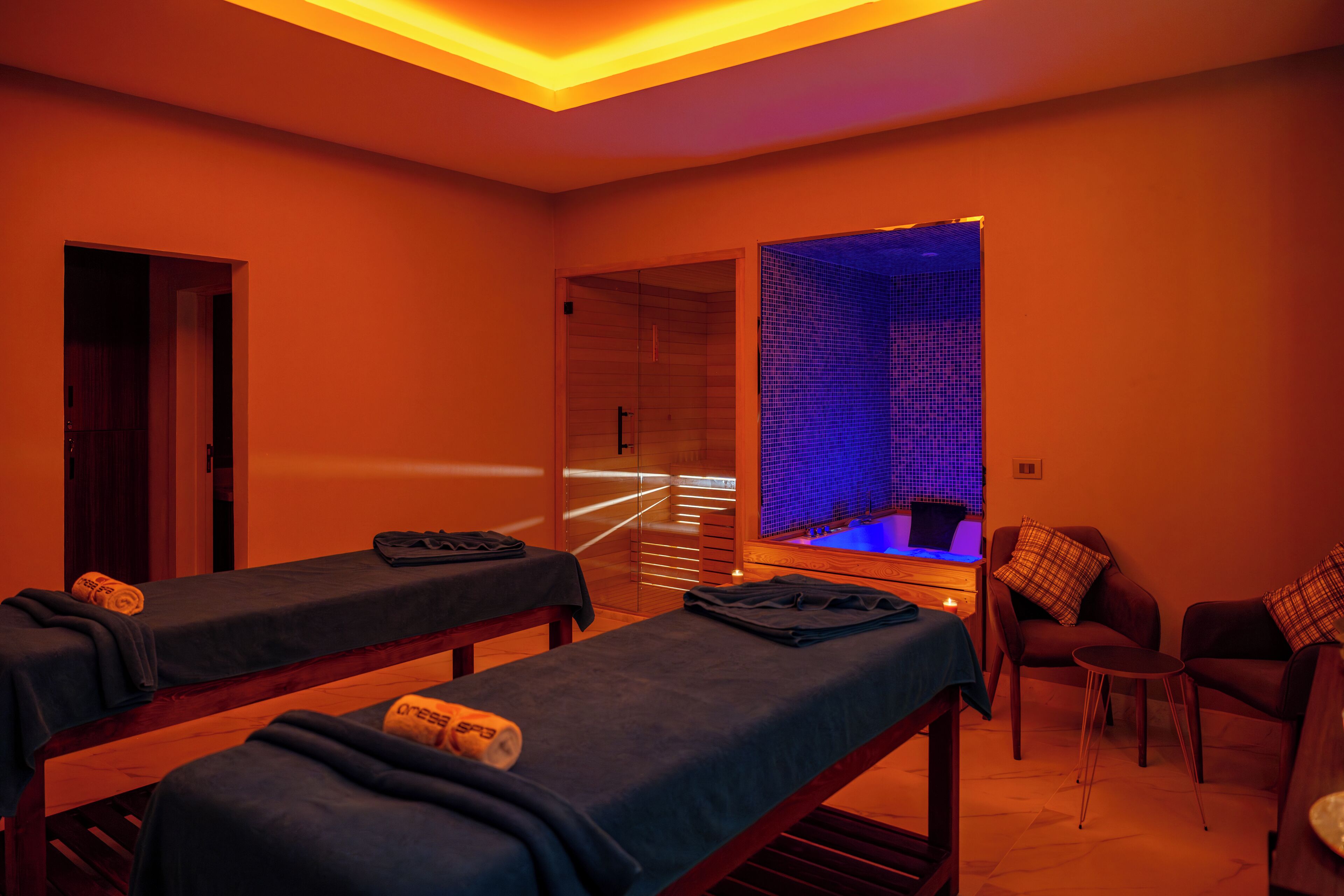 A spa room with two massage tables, a small seating area, and a blue-lit wall feature.