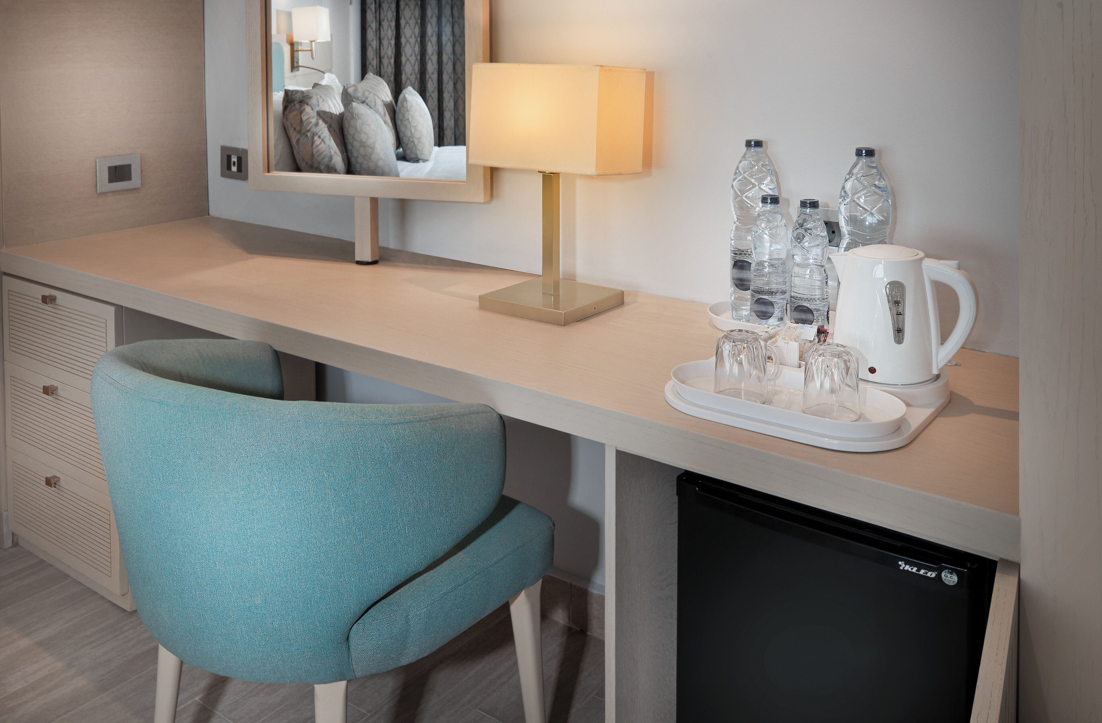 Une chambre d’hôtel avec un fauteuil de couleur turquoise, une table de chevet avec une lampe, des bouteilles d’eau et une machine à café.