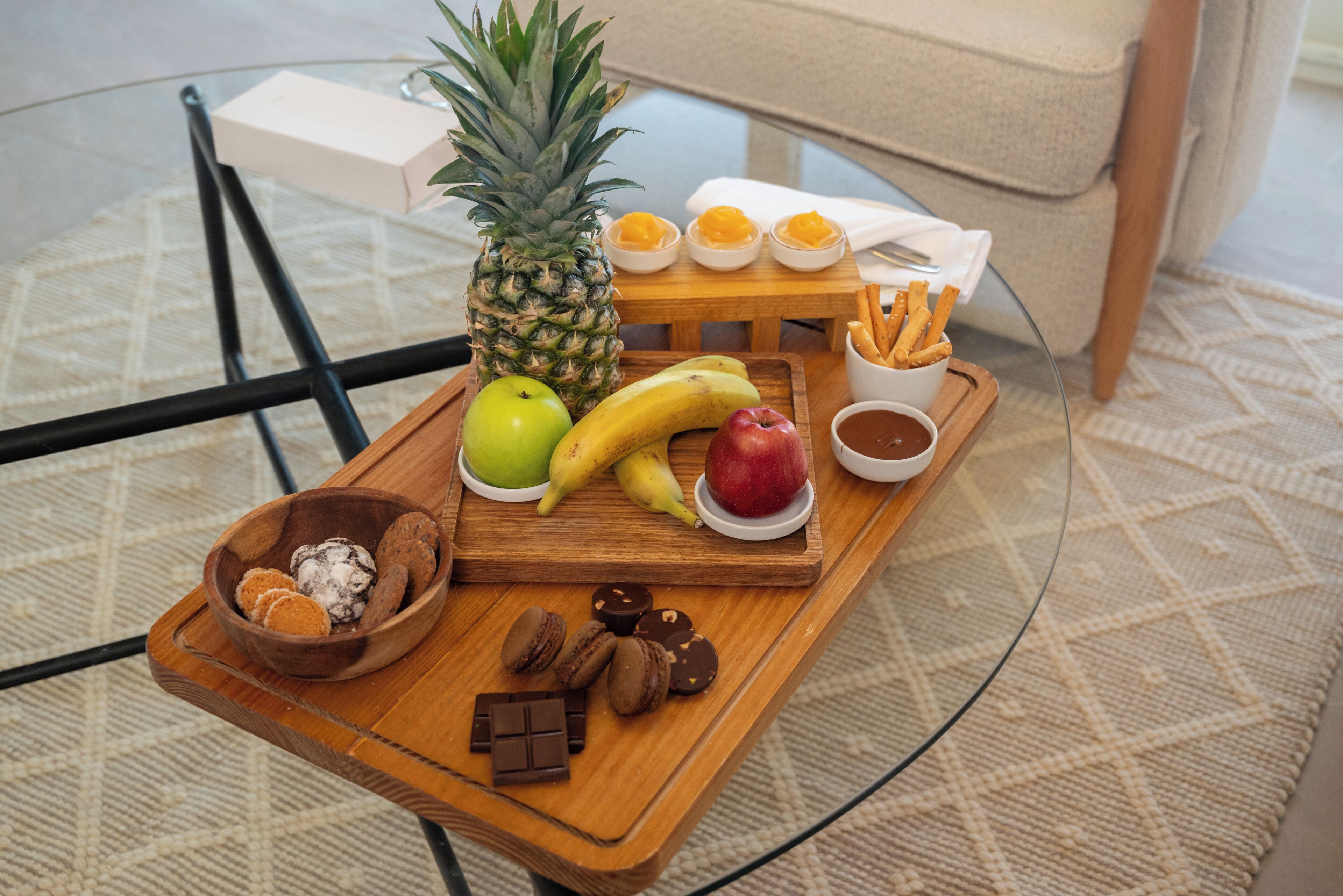A wooden tray with fruits, cookies, and chocolate on a glass table.