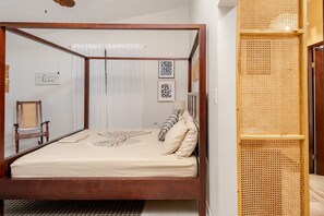 Un lit à baldaquin avec une structure en bois, un matelas blanc et des coussins à motifs. On y trouve également une chaise avec un siège en osier, un cadre photo fixé au mur et un paravent en osier.