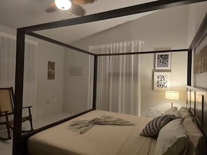 Una cama con dosel y cortinas, una mesita de noche con lámpara, y una silla.