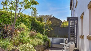 A patio area with a stone table, white chairs, and a wooden bench. There is a wooden fence and a variety of plants and shrubs. - 2 bedroom accommodation in Cark, near Grange-over-Sands (Cark, near Grange-over-Sands)