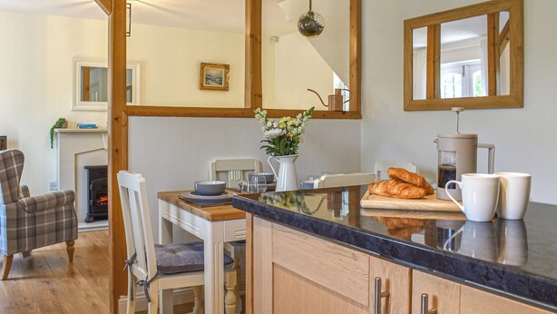 A kitchen with wooden cabinets, a marble countertop, a coffee maker, and a dining area with a chair and table. - 2 bedroom accommodation in Cark, near Grange-over-Sands (Cark, near Grange-over-Sands)