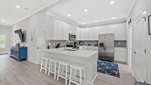 A modern kitchen with white cabinetry, a central island with bar stools, stainless steel appliances, and a blue patterned rug. - Castaway Cay - Beautiful 4 BR House with Pool (North Myrtle Beach)