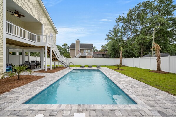 A rectangular swimming pool with a stone border, a covered patio with a ceiling fan, and a white fence surrounding the yard.