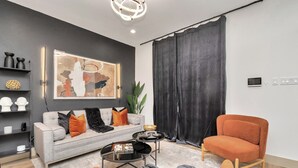 A modern living room with a grey sofa, orange armchair, and a coffee table. There is a large abstract painting on the wall, a plant, and a black curtain.