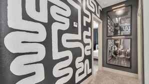 A hallway with a black and white abstract wall design, a framed mirror reflecting a room, and a carpeted floor.