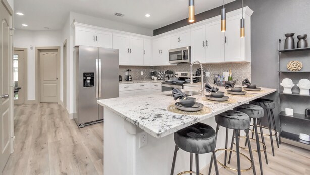 A modern kitchen with white cabinets, a large island with stools, stainless steel appliances, and pendant lighting.