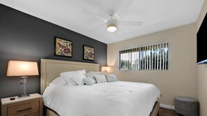 Ein Schlafzimmer mit Bett, Nachttisch, Lampe, Deckenventilator, einem Fenster mit Gitter und einem Bild an der Wand.