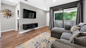 Ein modernes Wohnzimmer mit grauem Sofa, einem flachbildfernseher an der Wand, Holzboden und einem gemusterten Teppich.
