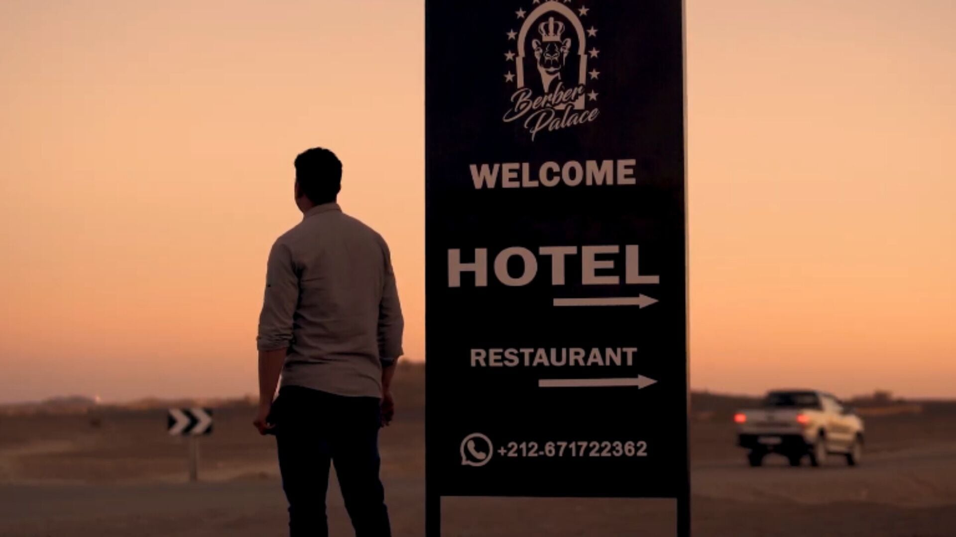 A man standing in front of a signboard with 'Welcome' and directional arrows to 'Hotel' and 'Restaurant' with a contact number.