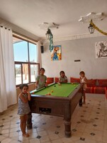 A pool table in a room with a tiled floor, a sofa with patterned cushions, and a painting on the wall.
