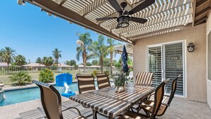 An outdoor patio with a table and chairs, a ceiling fan, and a view of a swimming pool and palm trees. - Desert Golf Getaway by GPM -Tennis+Pickleball+Priv. Pool+More! Walk to Festivals (Indio)