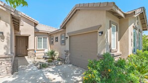 A two-story house with a beige exterior, a garage, and a small garden with plants and flowers. - Desert Golf Getaway by GPM -Tennis+Pickleball+Priv. Pool+More! Walk to Festivals (Indio)