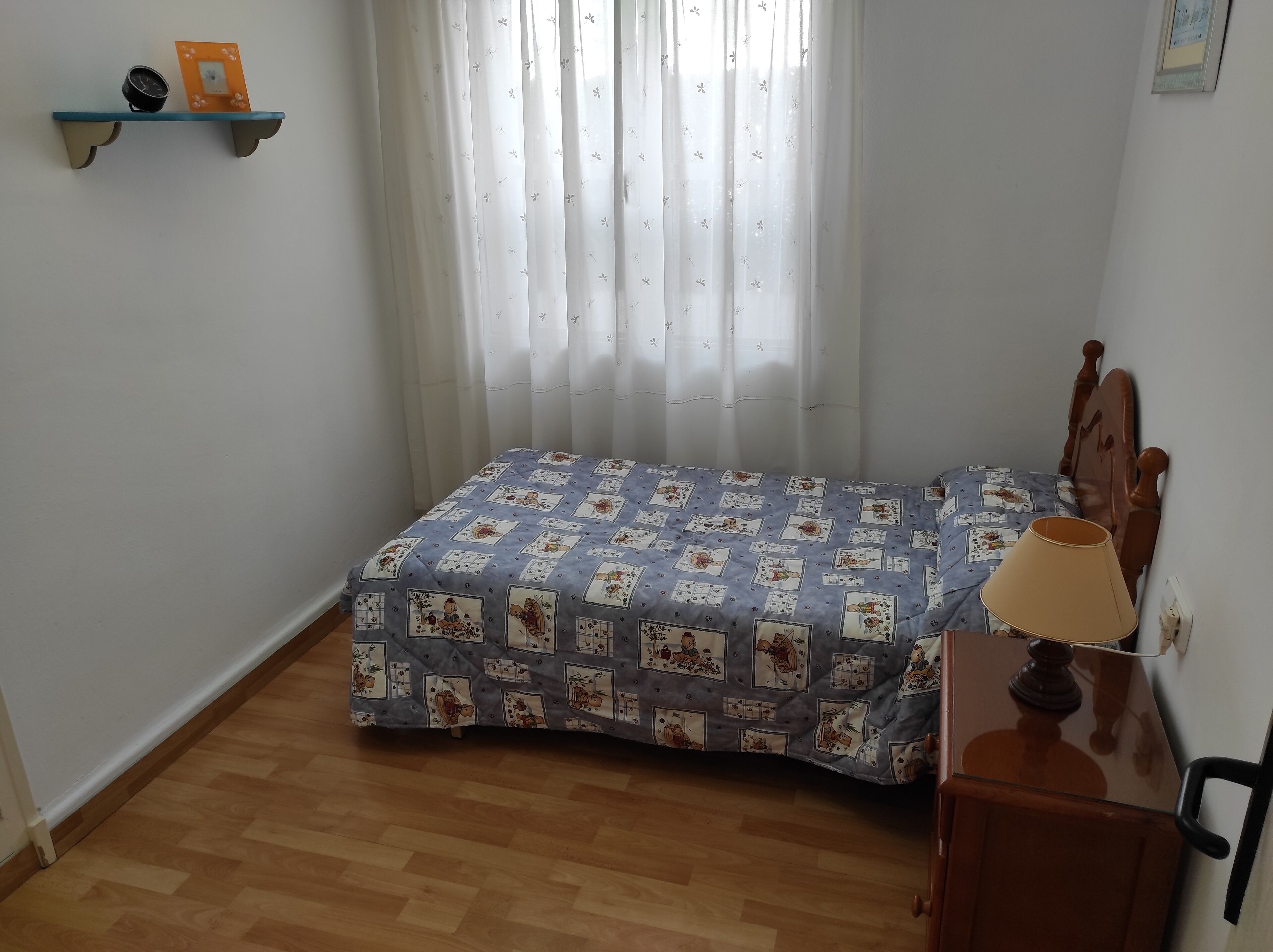 Ein kleines Schlafzimmer mit Bett, Nachttisch, Lampe, einer Kommode mit Uhr und Bild sowie einem Fenster mit durchscheinenden Vorhängen.