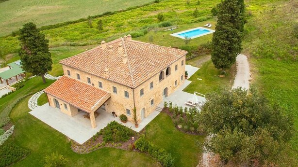 A large, two-story stone building with a tiled roof, surrounded by lush green fields and a swimming pool. - The Valley 3 (Castelfalfi)