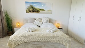 A neatly made bed with a white knitted blanket, two rolled towels, and a decorative woven placemat. There are two bedside lamps, a potted plant, and a framed picture on the wall. - Düne 77Modern vacation apartment in Westdorf in top location (Baltrum)