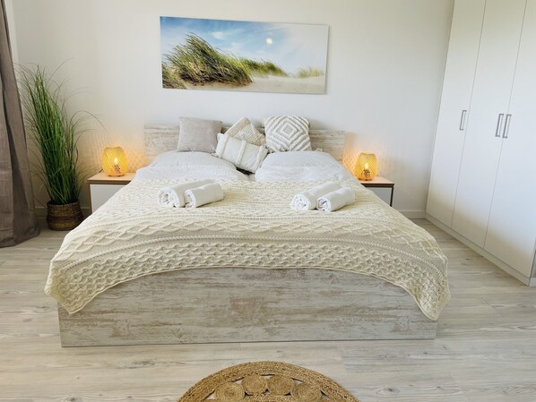 A neatly made bed with a white knitted blanket, two rolled towels, and a decorative woven placemat. There are two bedside lamps, a potted plant, and a framed picture on the wall. - Düne 77: Modern vacation apartment in Westdorf, ideally located (Baltrum)