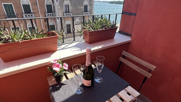 A balcony with a table set for two, featuring a bottle of wine and two glasses. There are potted plants on the balcony, and a view of a building and water in the background. - BIENNIAL PENTHOUSE WITH BEAUTIFUL PANORAMIC TERRACE OVER THE LAGOON FREE WI-FI (Venezia)