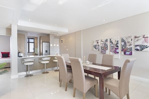 A modern dining area with a wooden table and beige chairs, a kitchen in the background, and a decorative butterfly wall art piece. - Eden on the Bay 256 (Cape Town)