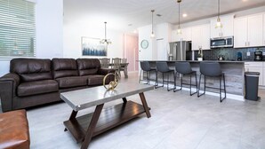 Una cocina moderna con armarios blancos, un refrigerador de acero inoxidable y un microondas. Hay un comedor con un sofá de cuero marrón y una mesa de centro.