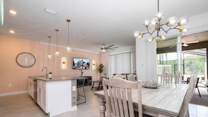 Una cocina amplia con una isla central, comedor con sillas de madera, ventilador de techo y un ventanal que proporciona luz natural.