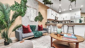 Un salon moderne avec un canapé gris, des coussins rouges et verts, et une table basse sur laquelle sont posés des livres. On aperçoit à l’arrière-plan une cuisine avec des meubles blancs et une plante suspendue.