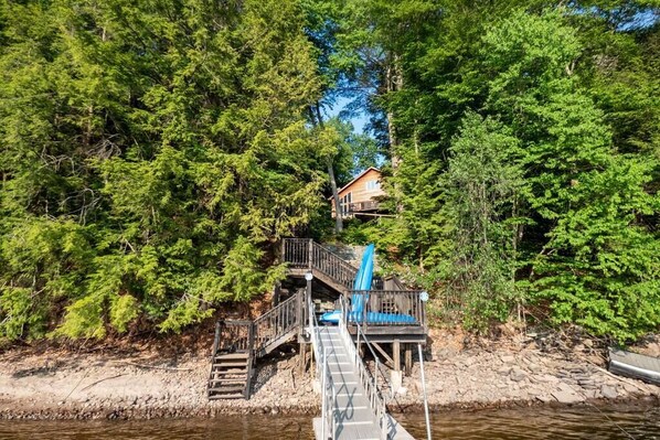 A cabin with a wooden deck and stairs leading to a blue chair, surrounded by dense green forest.
