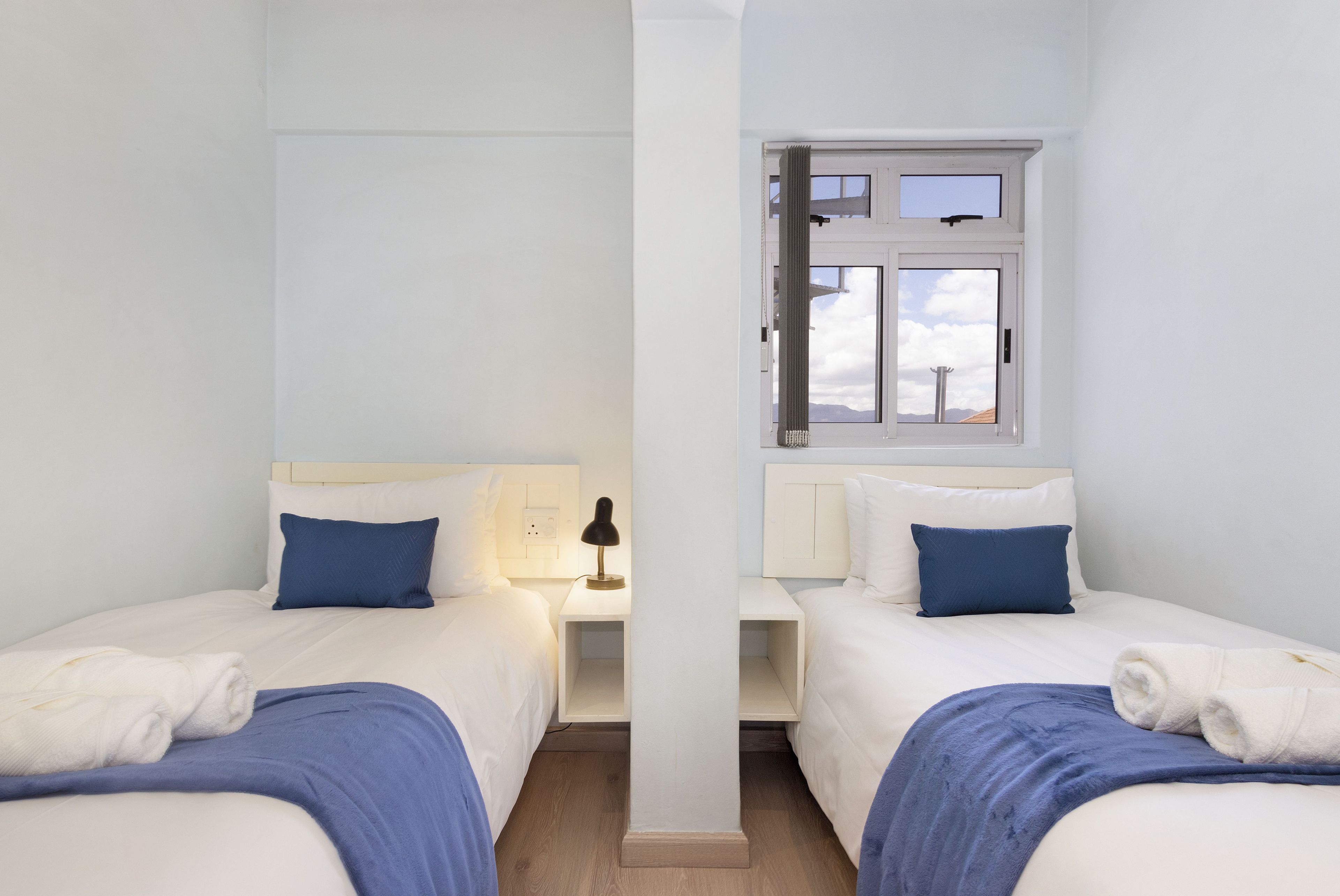 A hotel room with two beds, each with a white headboard, a nightstand, and a window with a view of the sky and mountains.