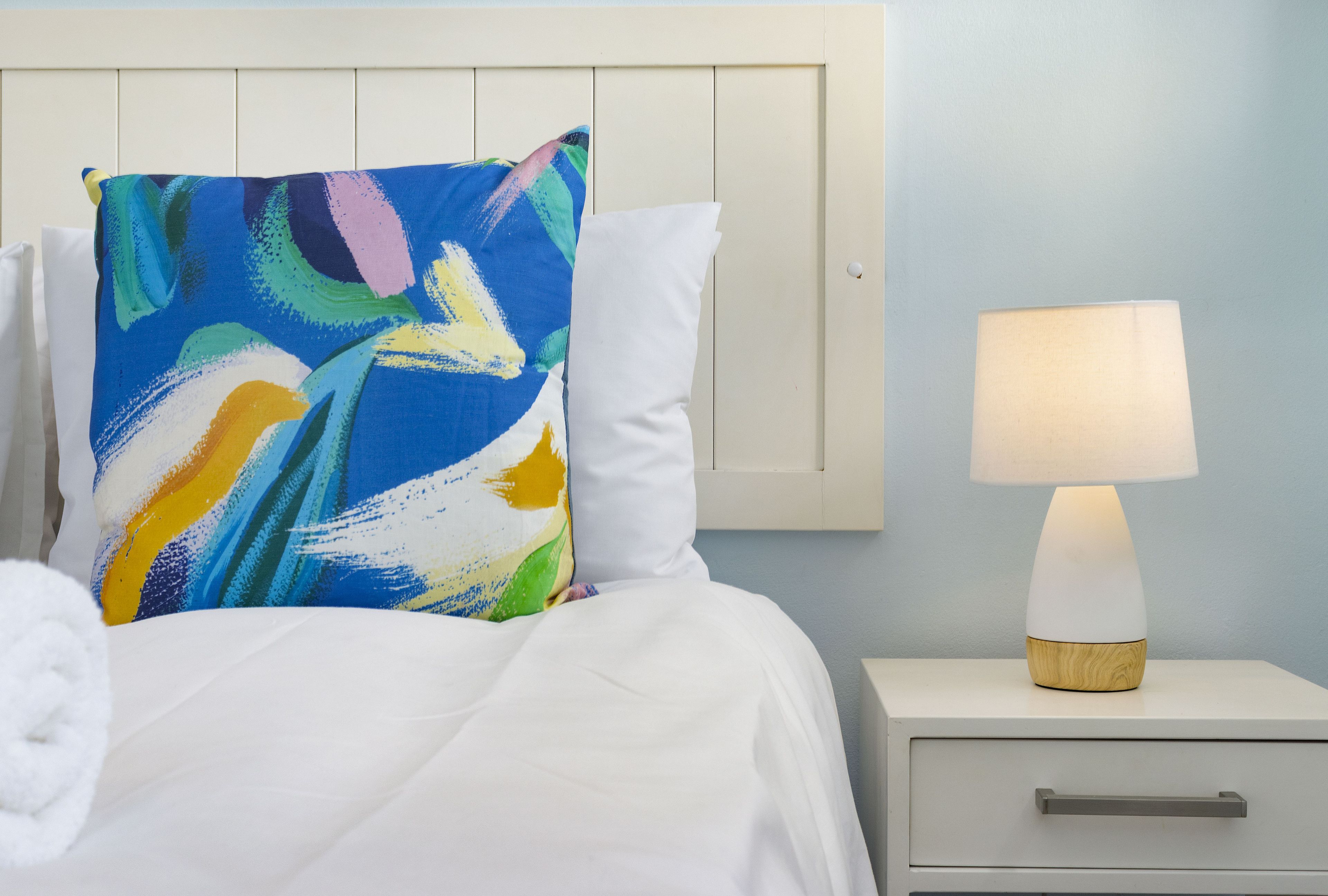 A neatly made bed with a colorful pillow and a white pillow, a bedside table with a lamp, and a drawer.