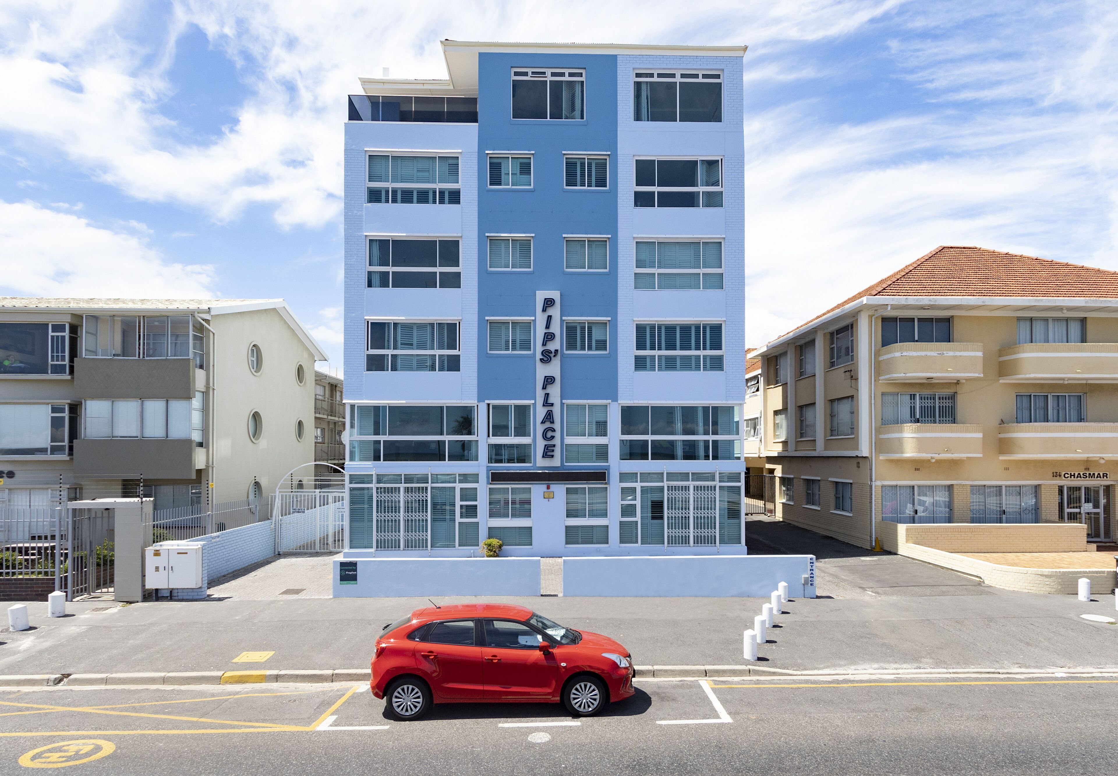 Un hotel de varios pisos con fachada moderna, un letrero llamativo y un estacionamiento con un auto rojo.