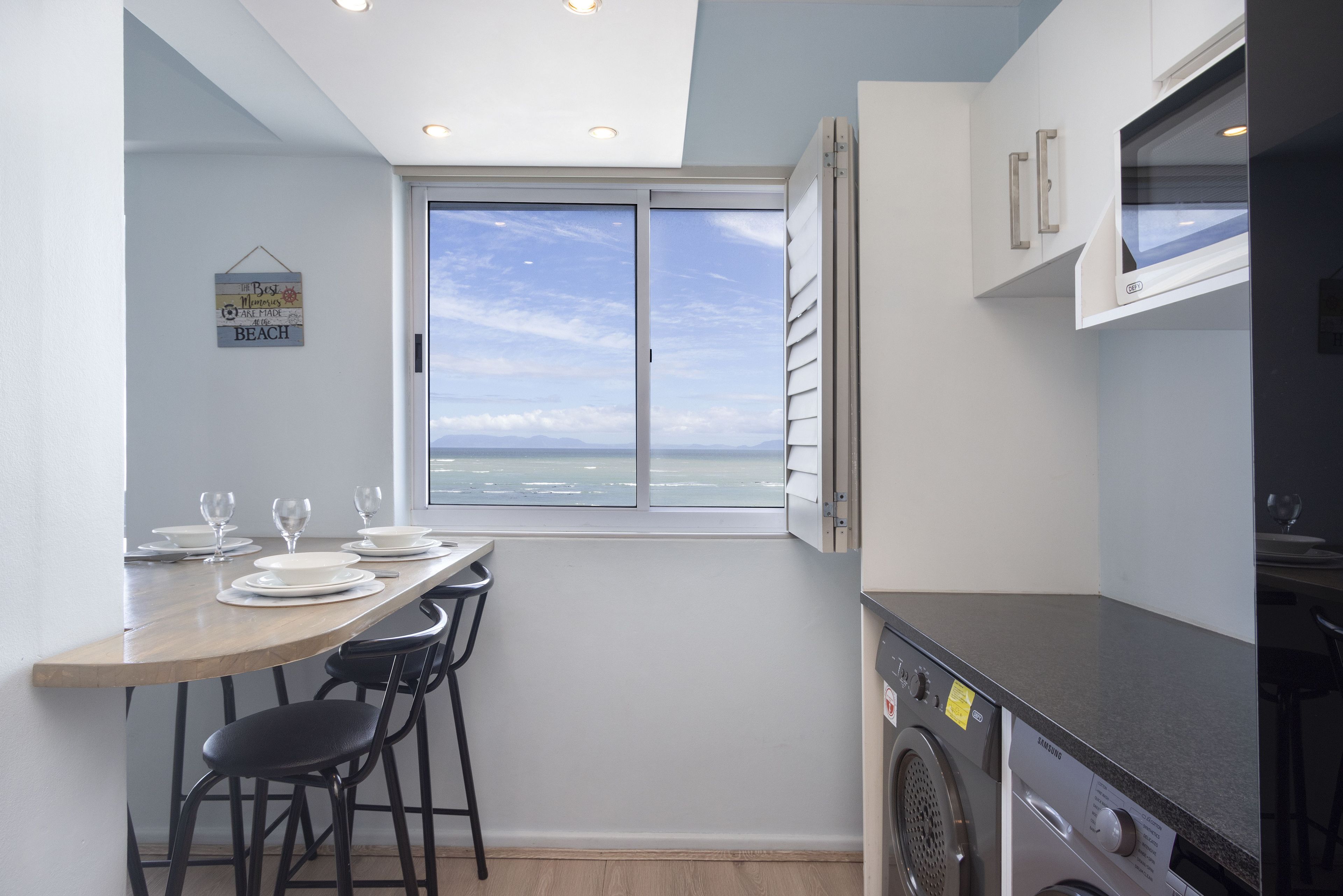A compact kitchen with a large window, a washing machine, and a dining area with chairs and plates.