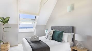 A bedroom with a bed, pillows, a bedside table with a lamp, a window, and a plant.