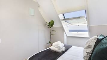 A bedroom with a skylight, a bed with a dark blanket, a plant, and a view of the sky.