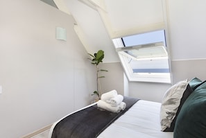 A bedroom with a skylight, a bed with a dark blanket, a plant, and a view of the sky. - Dolphin Beach H301 (Cape Town)