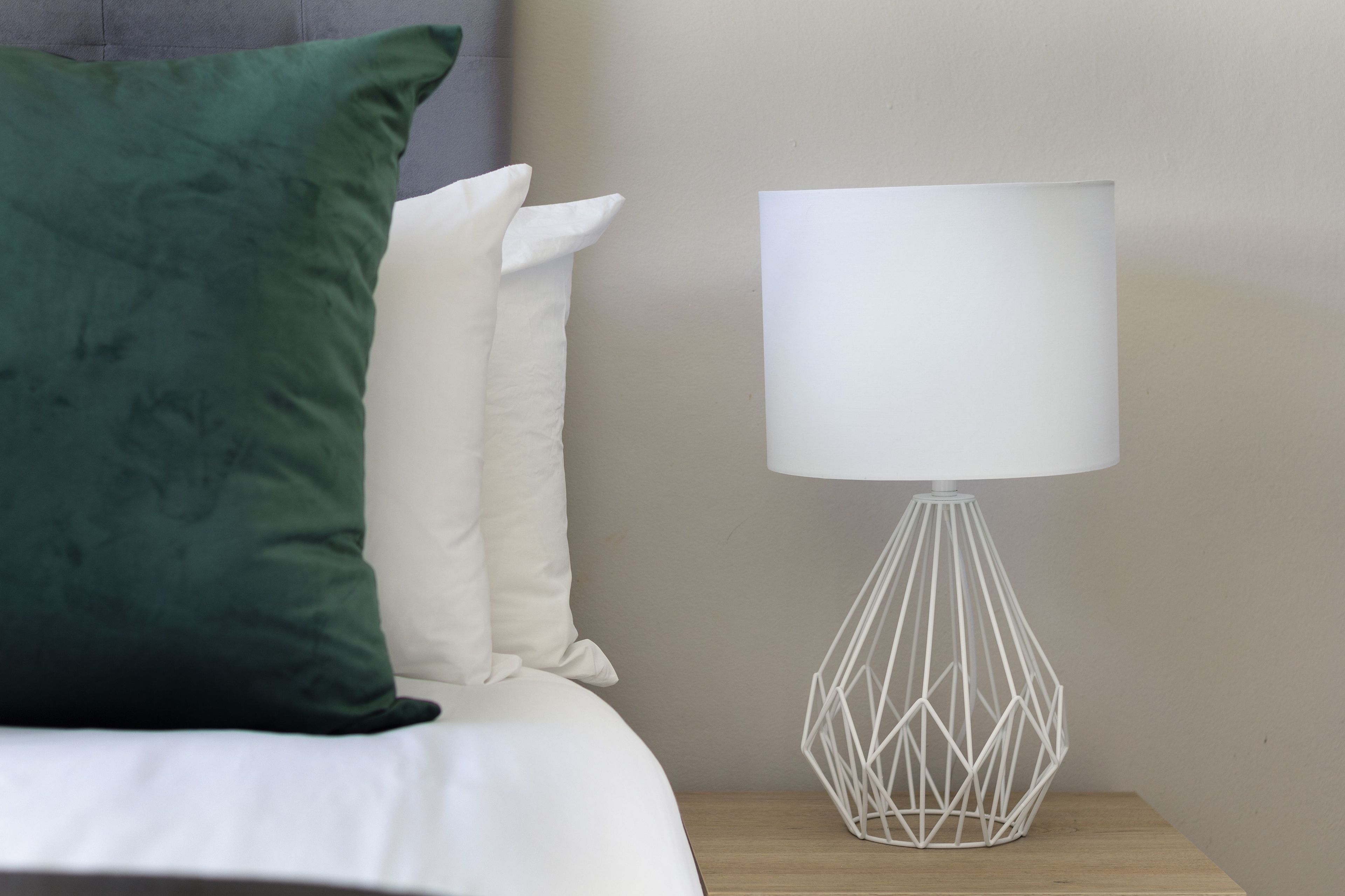 A bed with white and green pillows, a white lamp with a geometric base, and a wooden nightstand.
